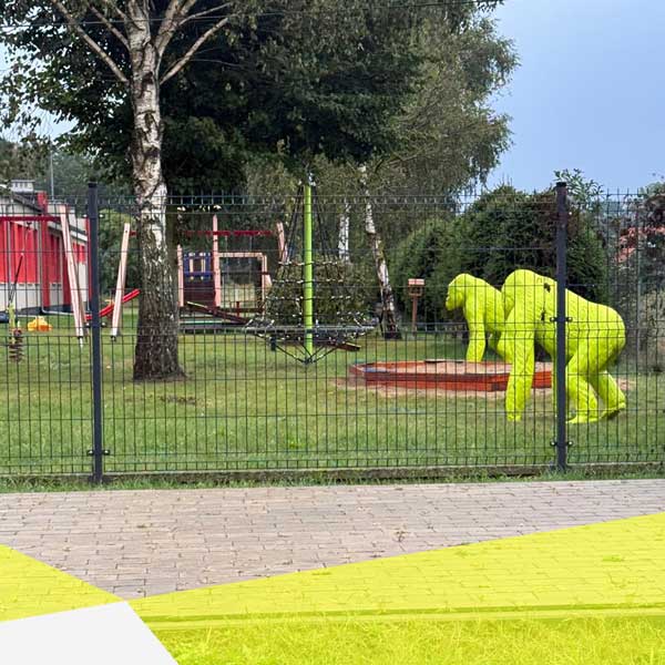 Hinter einem grünen Drahtzaun sieht man eine Außenanlage eines Kindergarten mit zwei grünen Gorillas, Sandkiste, Klettergerüsten und Schaukeln.