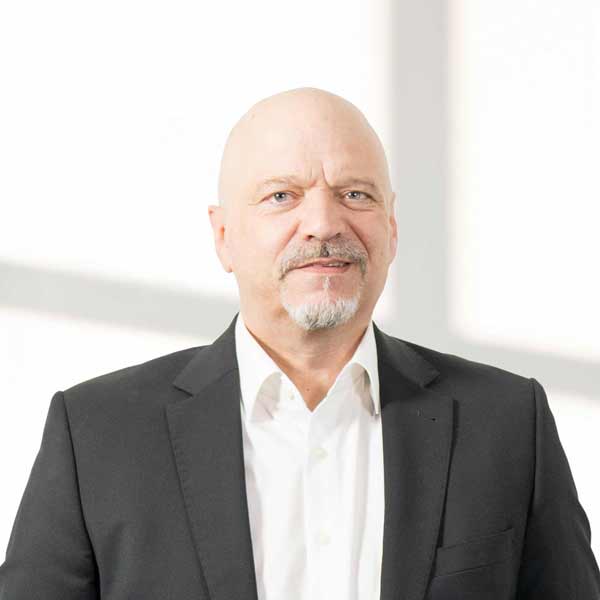 Ingo Burkhardt steht in einem weißen Raum, das Sonnenlicht scheint herein und man sieht die Schatten des Fensterrahms. Er steht frontal zur Kamera. Ist ab dem Oberkörper angeschnitten. Er trägt ein weißes Hemd und ein schwarzes Sacko. Er hat eine Glatze, ein grauen Oberlippen Bart und vorne einen kurzen Bart. Er grinst leicht in die Kamera.